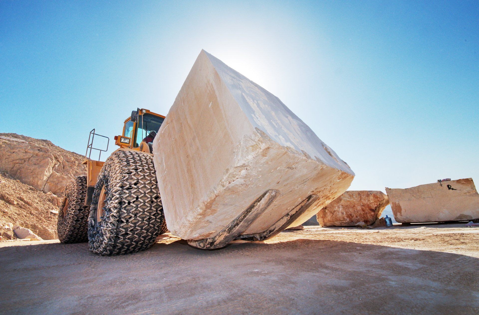 Front loader carrying a large, light-colored square stone block in a quarry, under a bright blue sky.