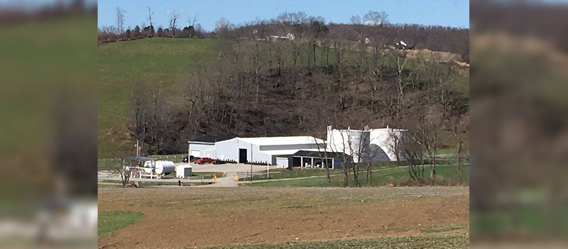 A white industrial building with large circular silos sits in a rural field with a wooded hillside in the background.
