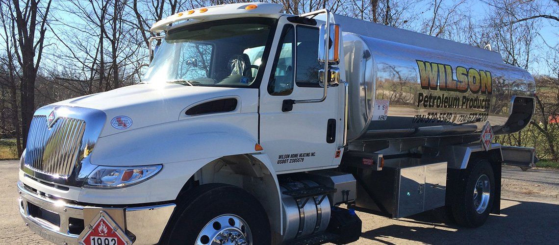 A white Wilson Petroleum tanker truck parked on a gravel surface outdoors with a backdrop of trees.