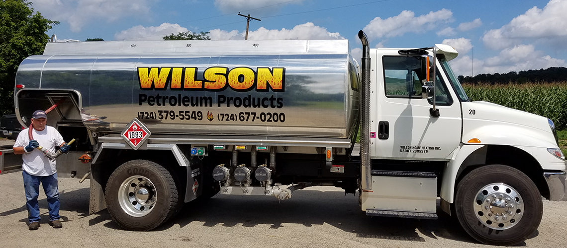 A person stands beside a parked Wilson Petroleum Products fuel tanker truck in a gravel lot next to a cornfield.