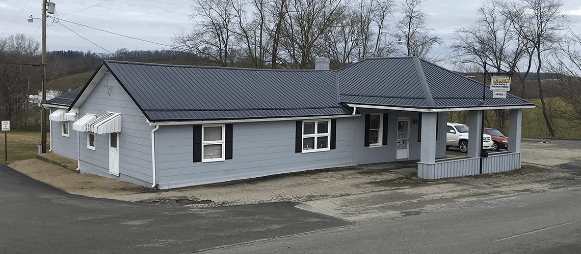 A gray, single-story building with a dark metal roof and black window shutters sits near a road under a cloudy sky.