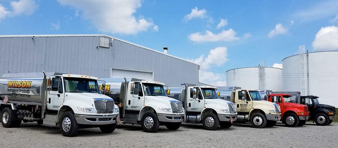 A line of tanker trucks parked in front of a metal warehouse and large white silos under a blue sky with clouds.