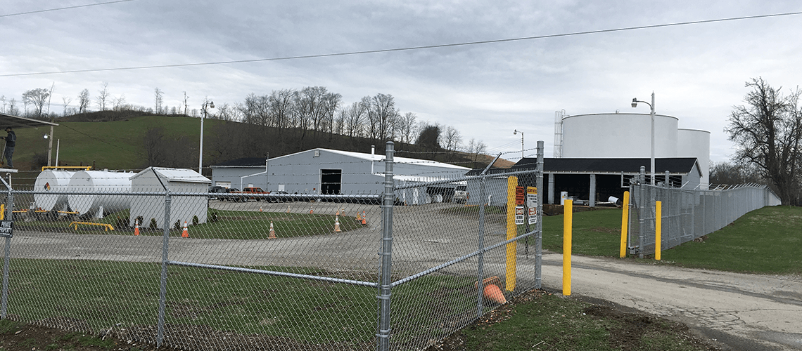 An industrial site with storage tanks and warehouses behind a chain-link fence under a cloudy sky.