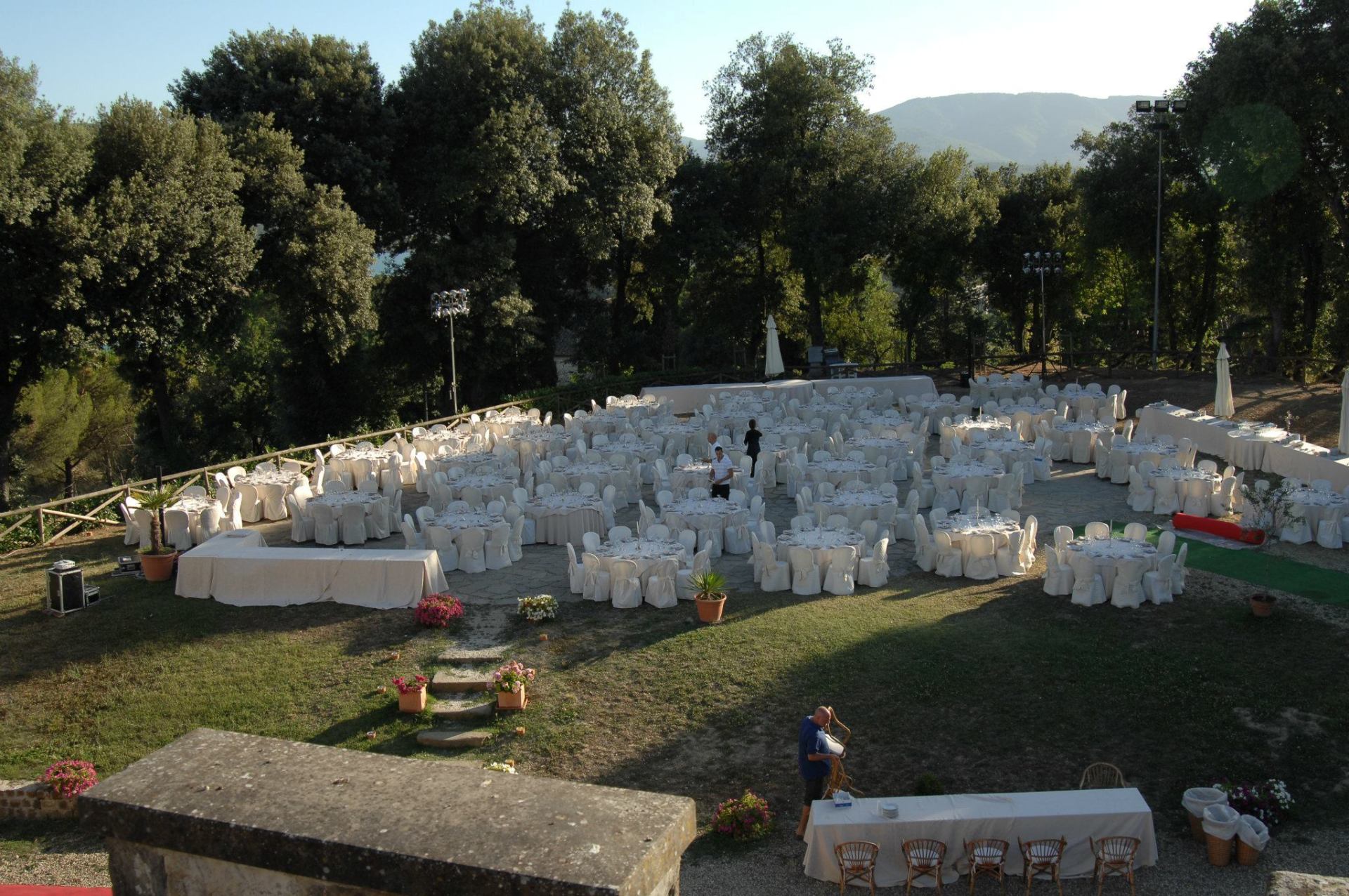 Banchetto all'aperto con numerose sedie ricoperte di bianco e tavoli rotondi allestiti di fronte a un castello di pietra al tramonto.