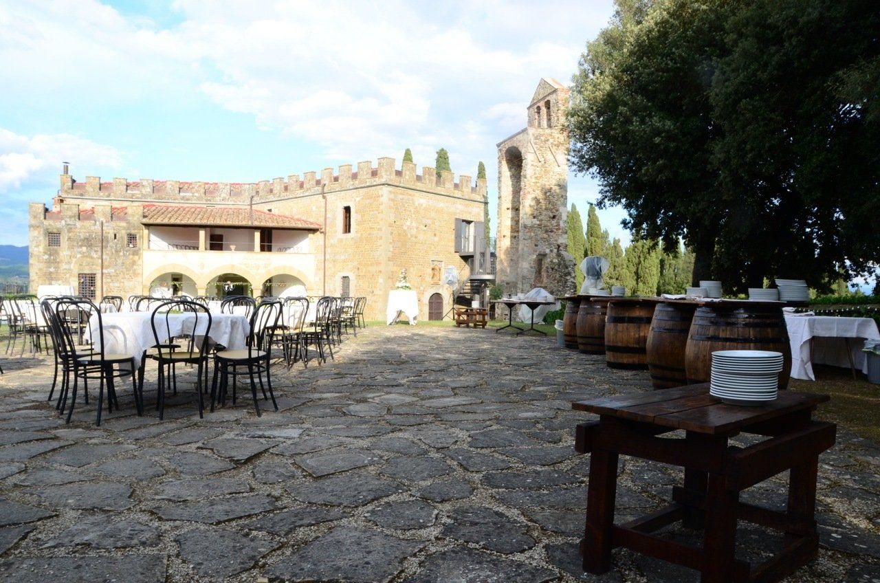 Cortile in pietra con tavolini e sedie da caffè accanto a un edificio storico simile a un castello e ad alberi.