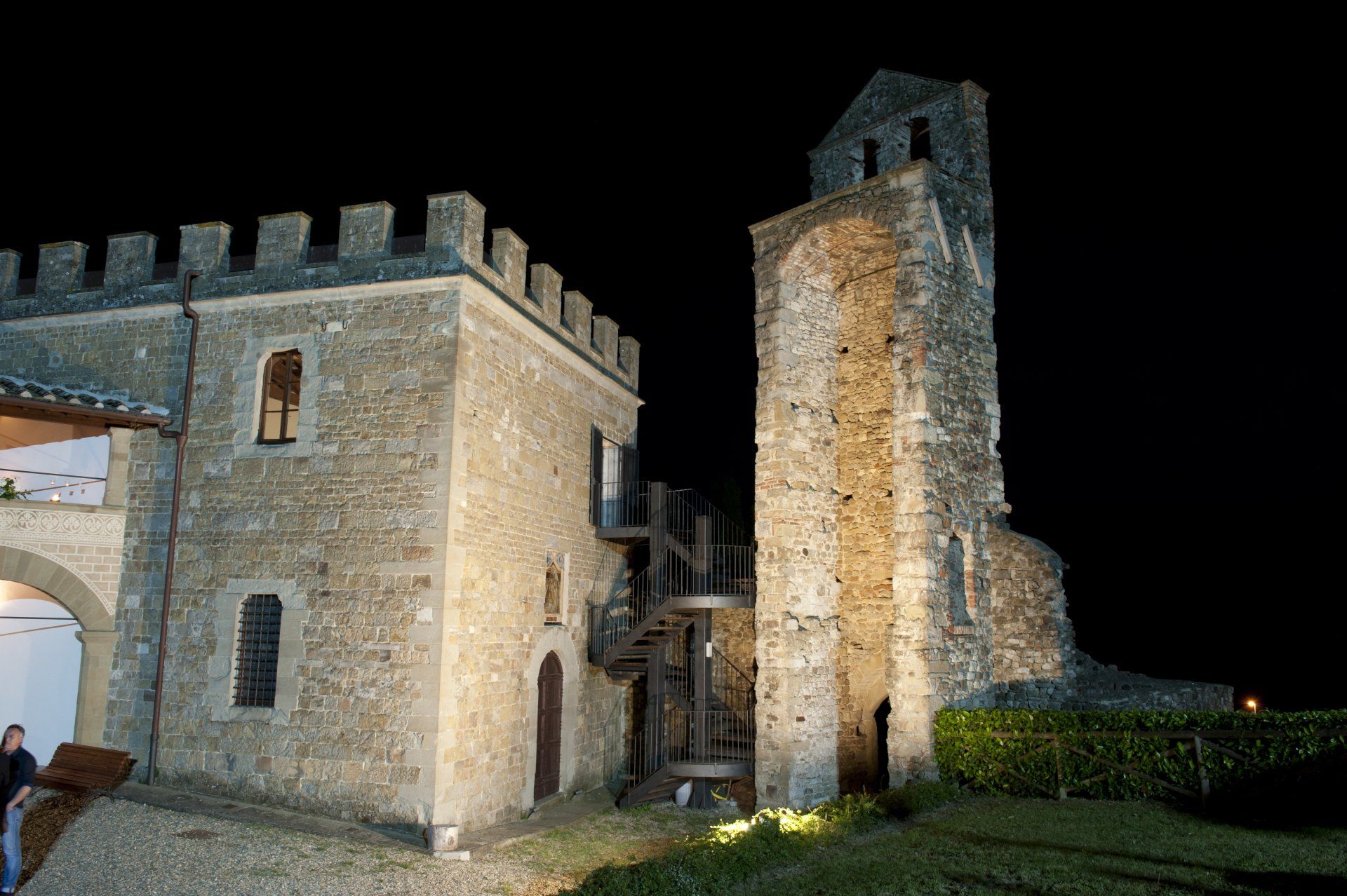 Torre e merlature di un castello in pietra illuminate di notte, stagliate contro un cielo nero.