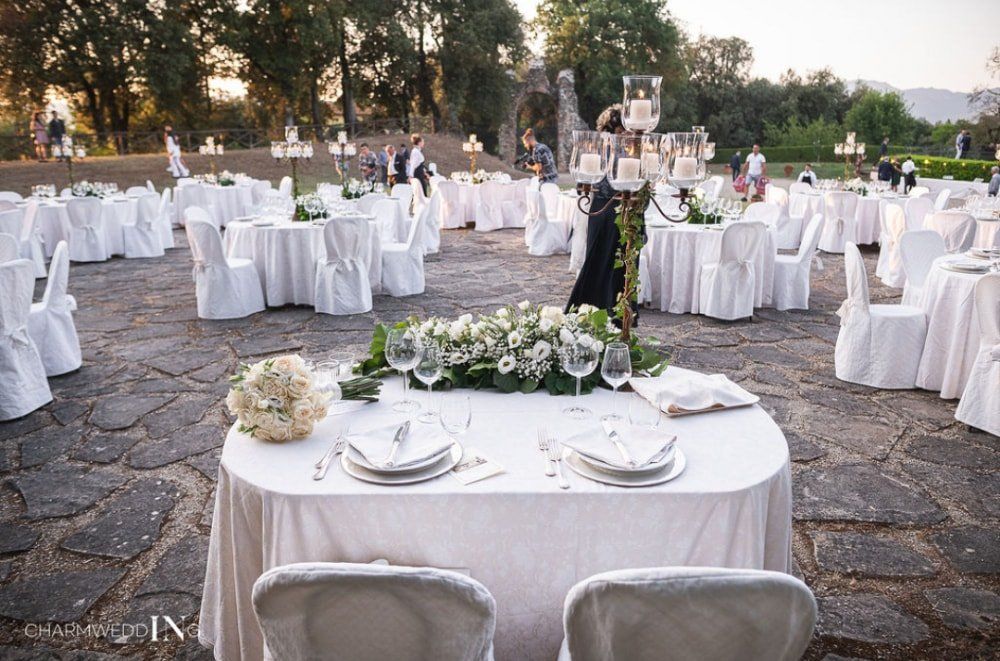 Elegante ricevimento di nozze all'aperto con tavoli apparecchiati con drappeggi bianchi, centrotavola floreali e un'ambientazione in un cortile di pietra.
