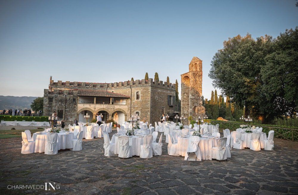 Ricevimento di nozze all'aperto con tavoli apparecchiati con tovaglie bianche in un cortile in pietra accanto a un castello storico al tramonto.