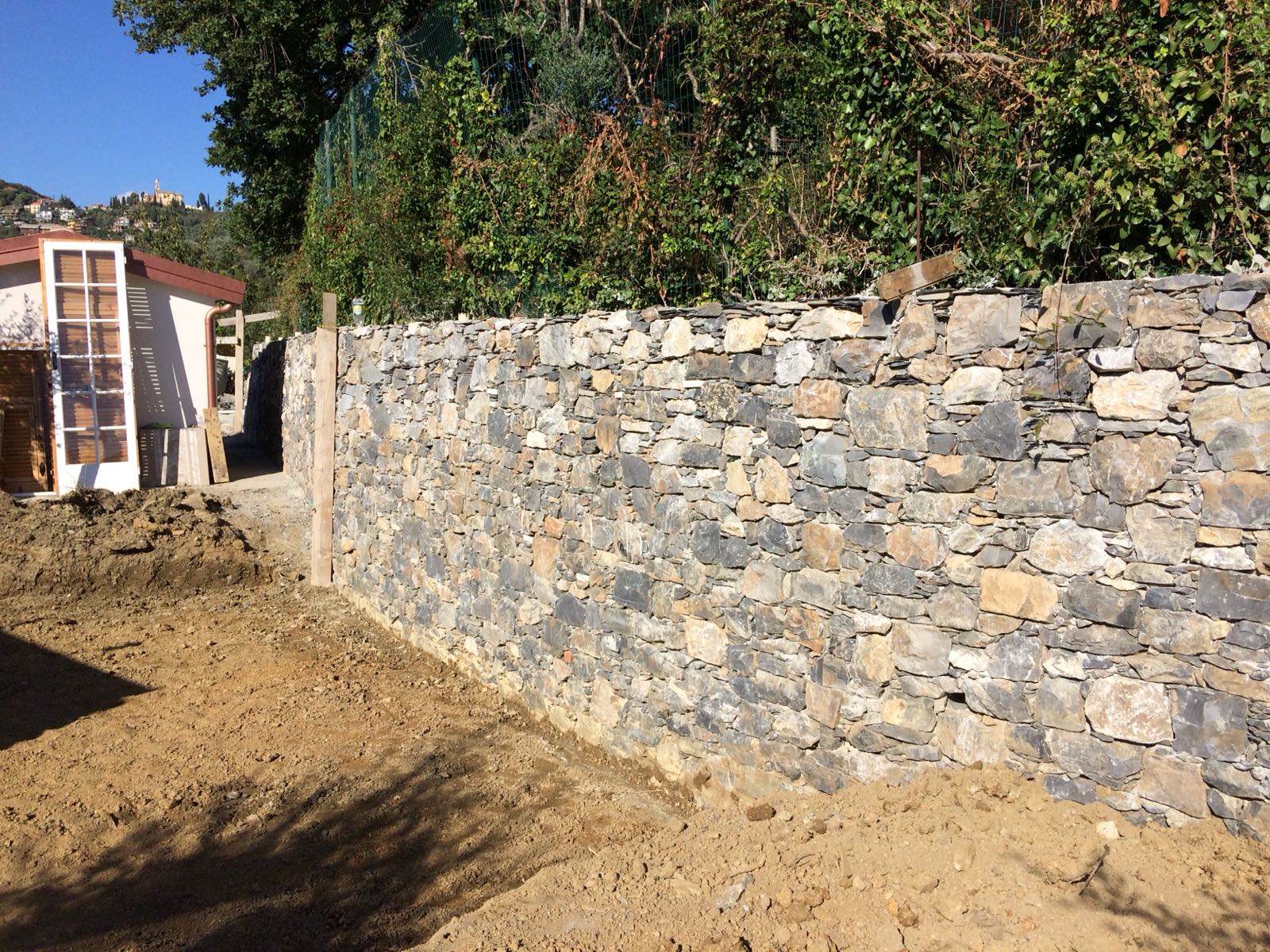 Muro di pietra in costruzione con terreno sterrato davanti e fogliame sullo sfondo.