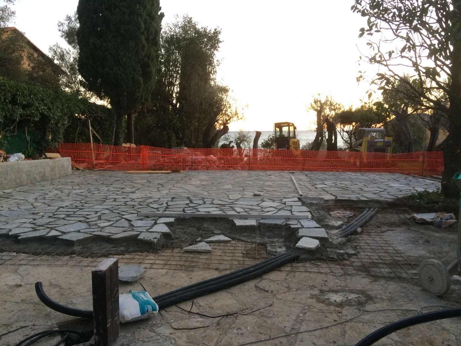 Cantiere: pavimentazione in ciottoli in corso, recinzione di sicurezza arancione, macchinari pesanti sullo sfondo vicino all'acqua.