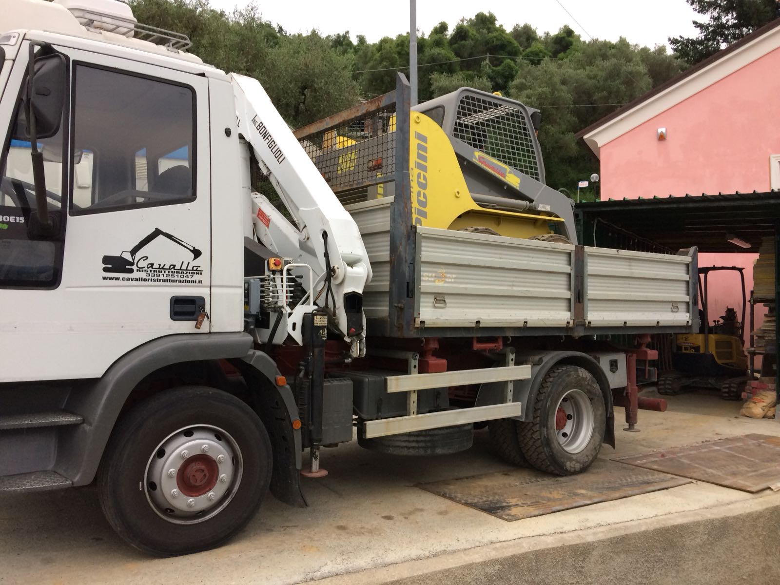 Camion bianco con una gru che trasporta una macchina edile gialla.