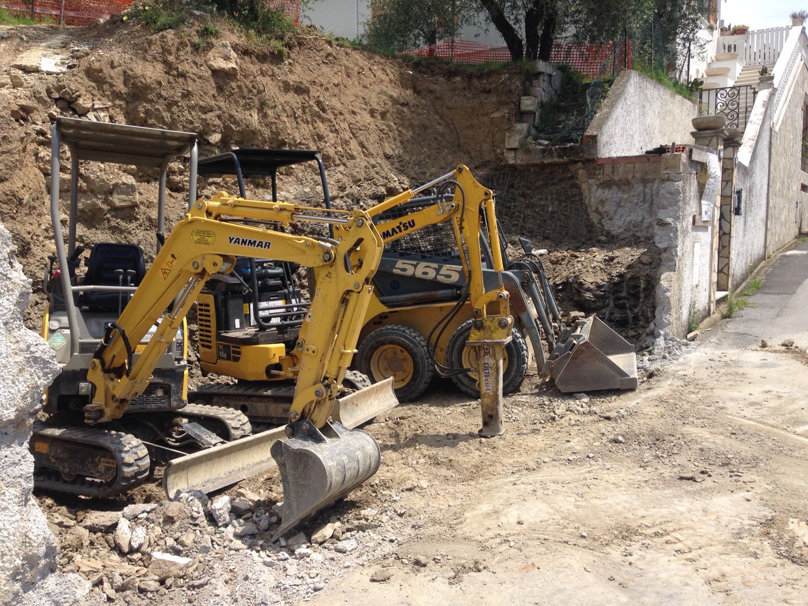 Escavatore giallo e minipala al lavoro in un cantiere edile accanto a un muro di pietra.