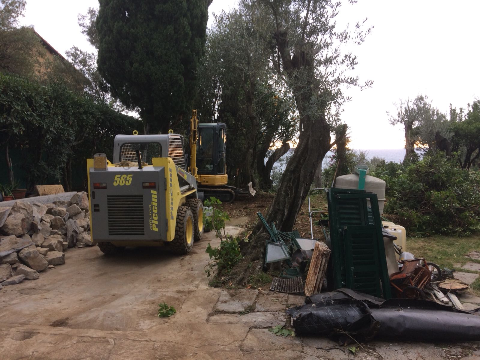 Una minipala e un escavatore lavorano all'aperto su un sentiero di pietra, con alberi e oceano sullo sfondo.