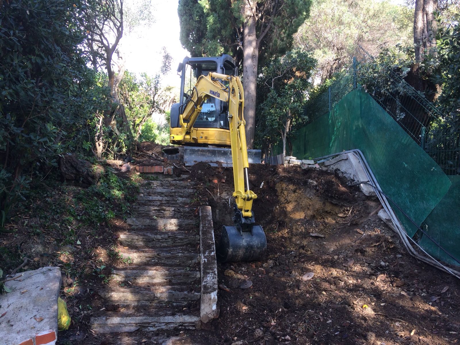 Escavatore giallo sulle scale, impegnato a rimuovere terra e detriti. Contesto boschivo, recinzione verde sul lato destro.