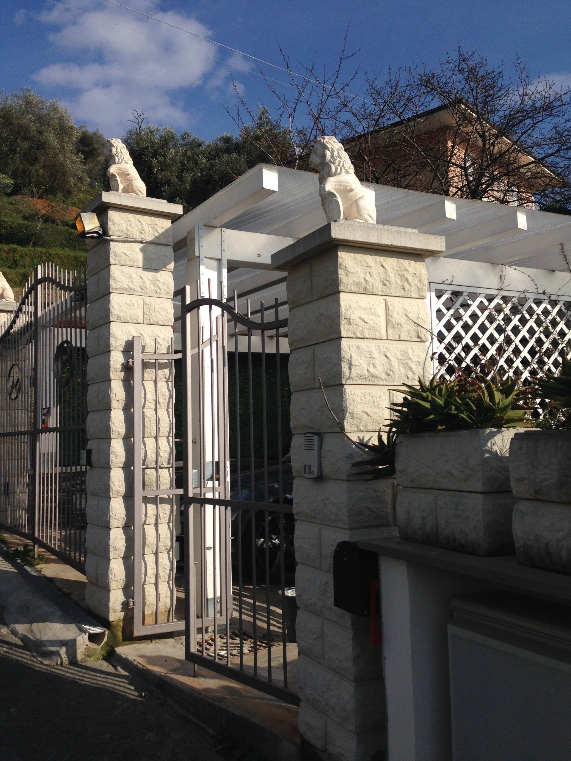 Ingresso con cancello in pietra, cancelli in ferro, sculture di leoni in cima ai pali e un pergolato bianco.