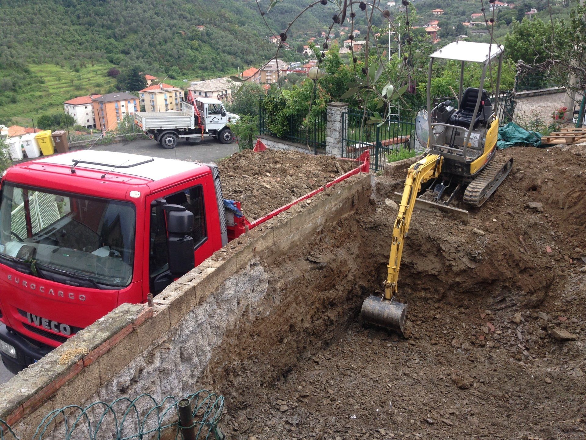 Cantiere con un camion rosso e un piccolo escavatore che scavano in una zona collinare.