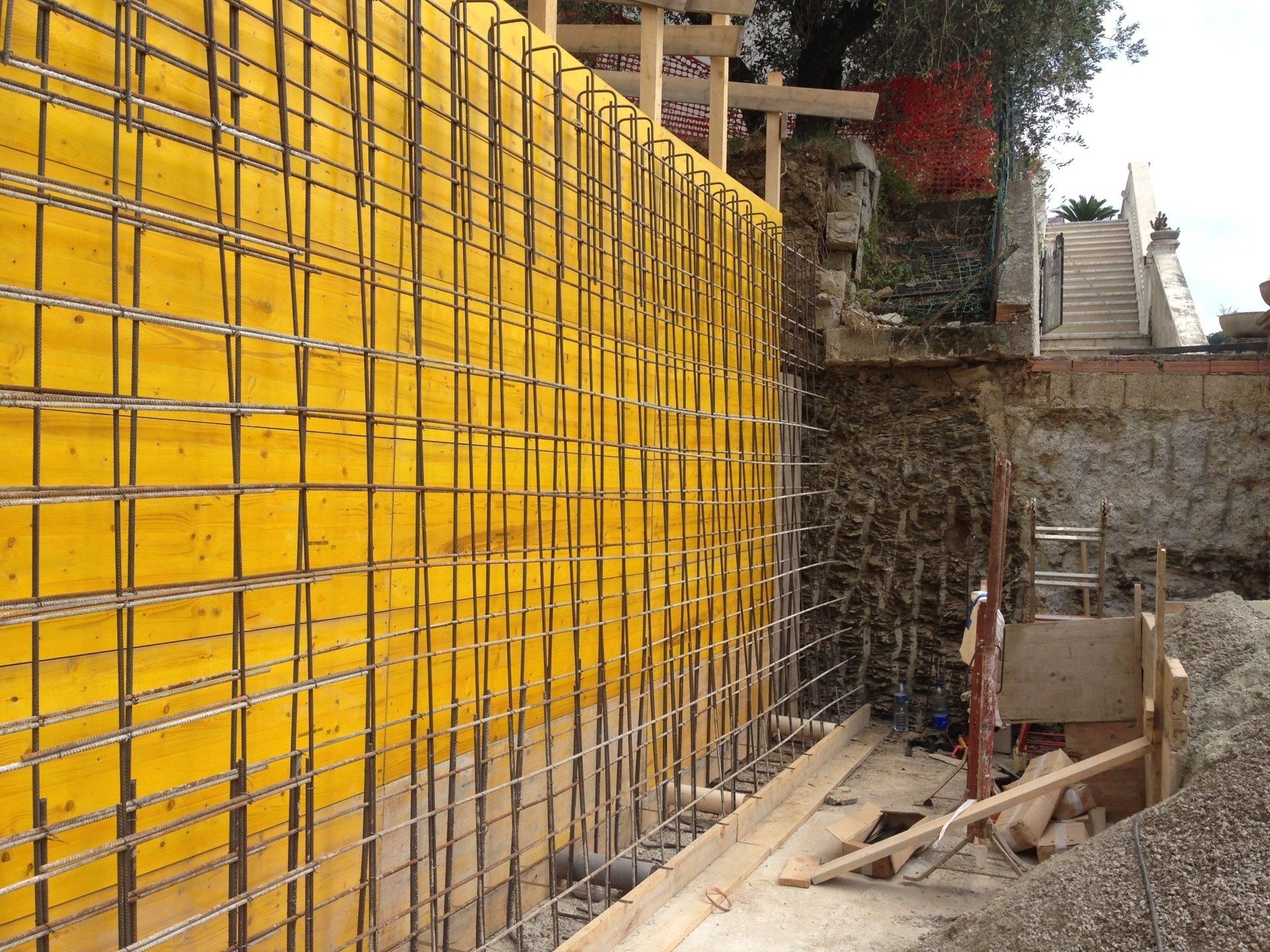 Cantiere con casseforme gialle, barre d'armatura e muro di contenimento in cemento parzialmente costruito.