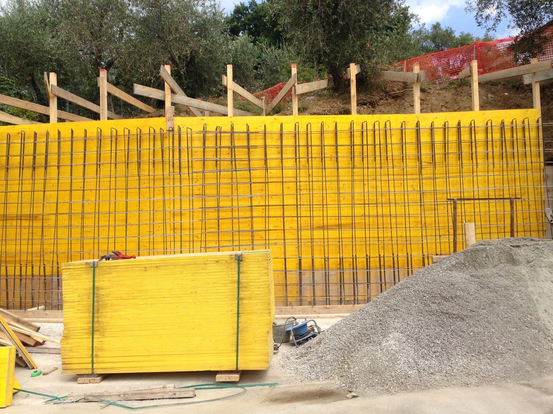 Cantiere: cassero in cemento giallo, griglia di tondini di armatura, cumulo di ghiaia e supporti in legno.