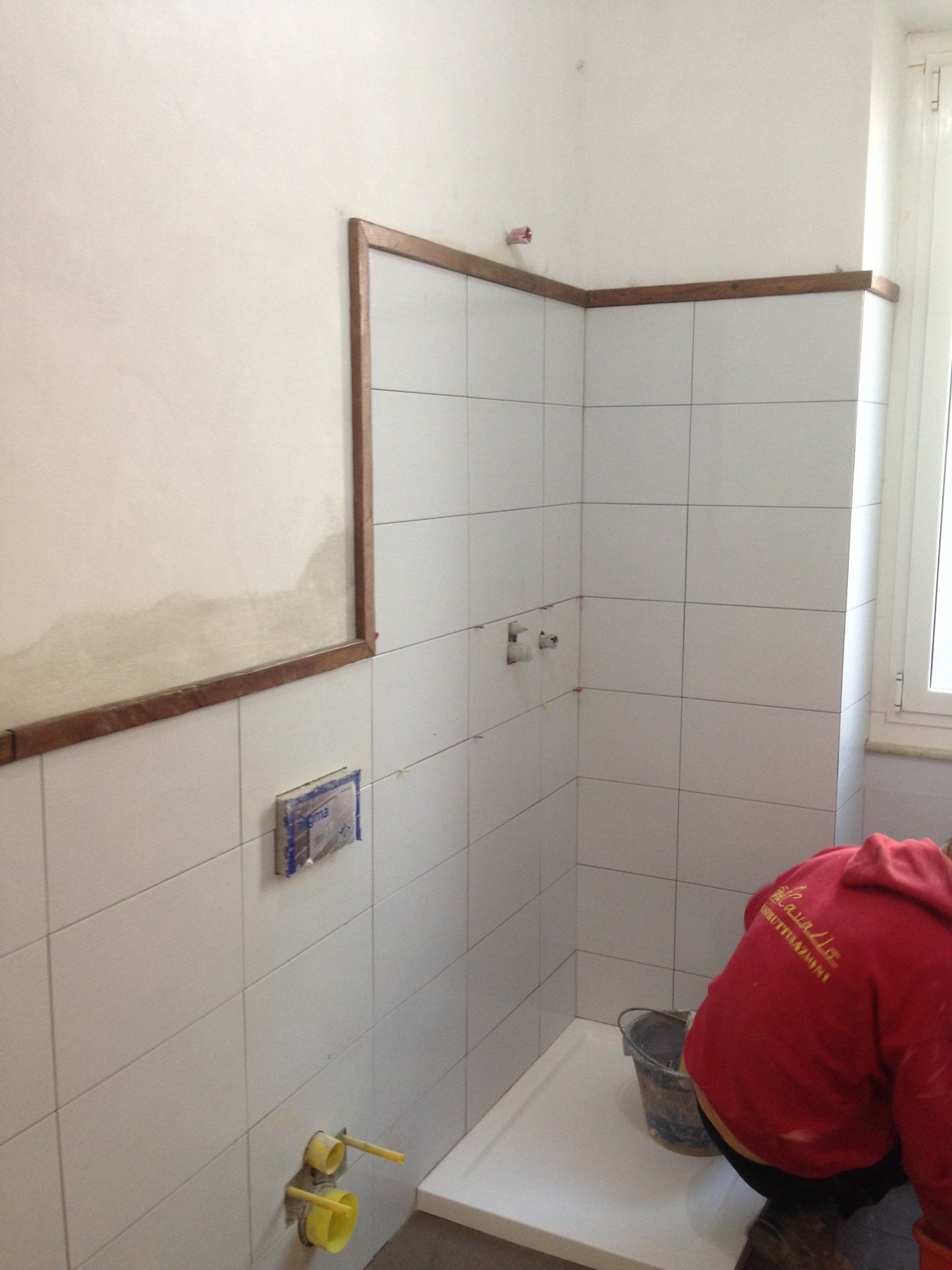 Bagno in costruzione con piastrelle bianche, finiture marroni e una persona con una camicia rossa al lavoro.