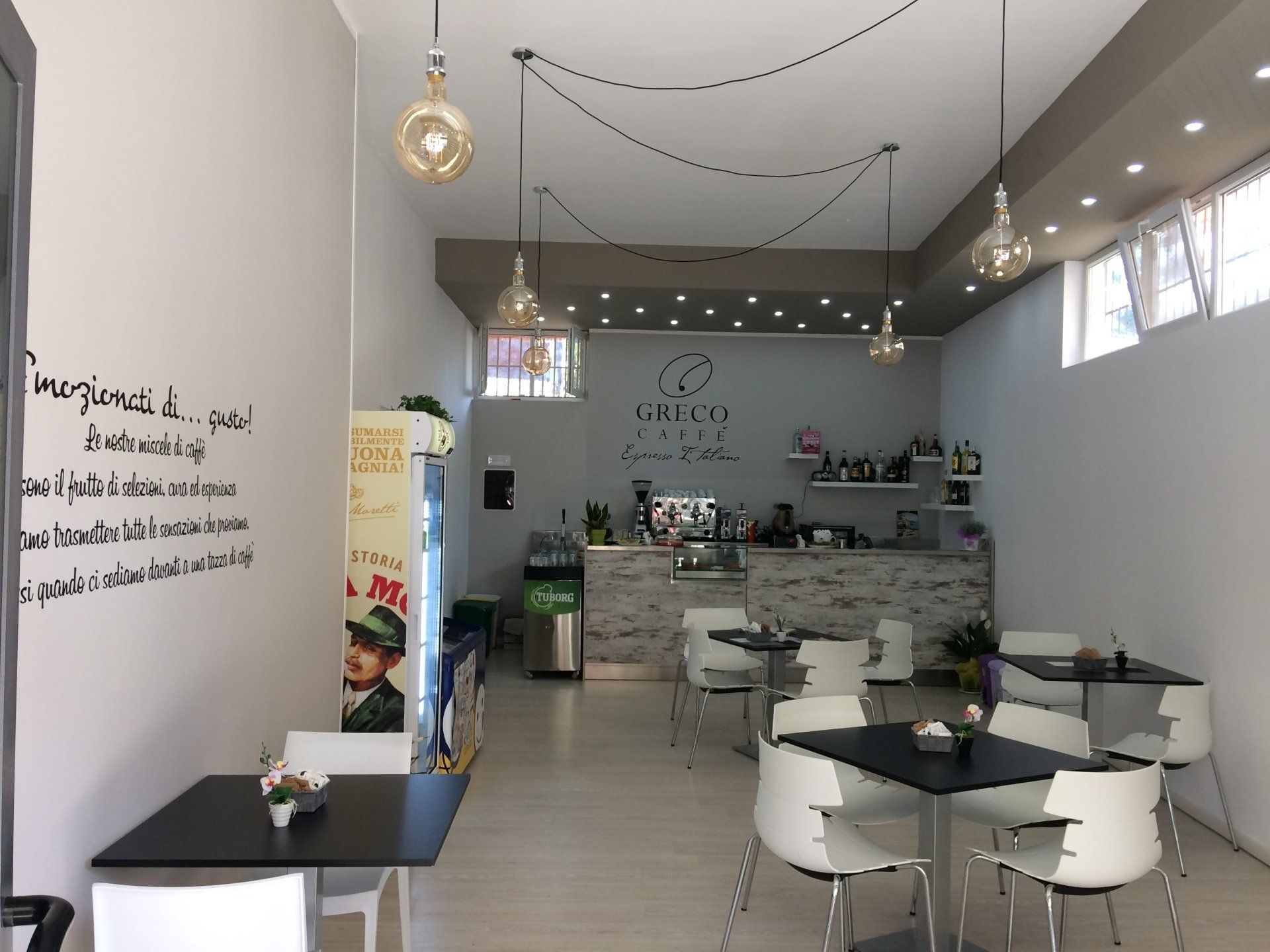 Interno di un bar con tavoli, sedie, bancone e luci decorative sospese. È visibile un frigorifero con la pubblicità di una bevanda.