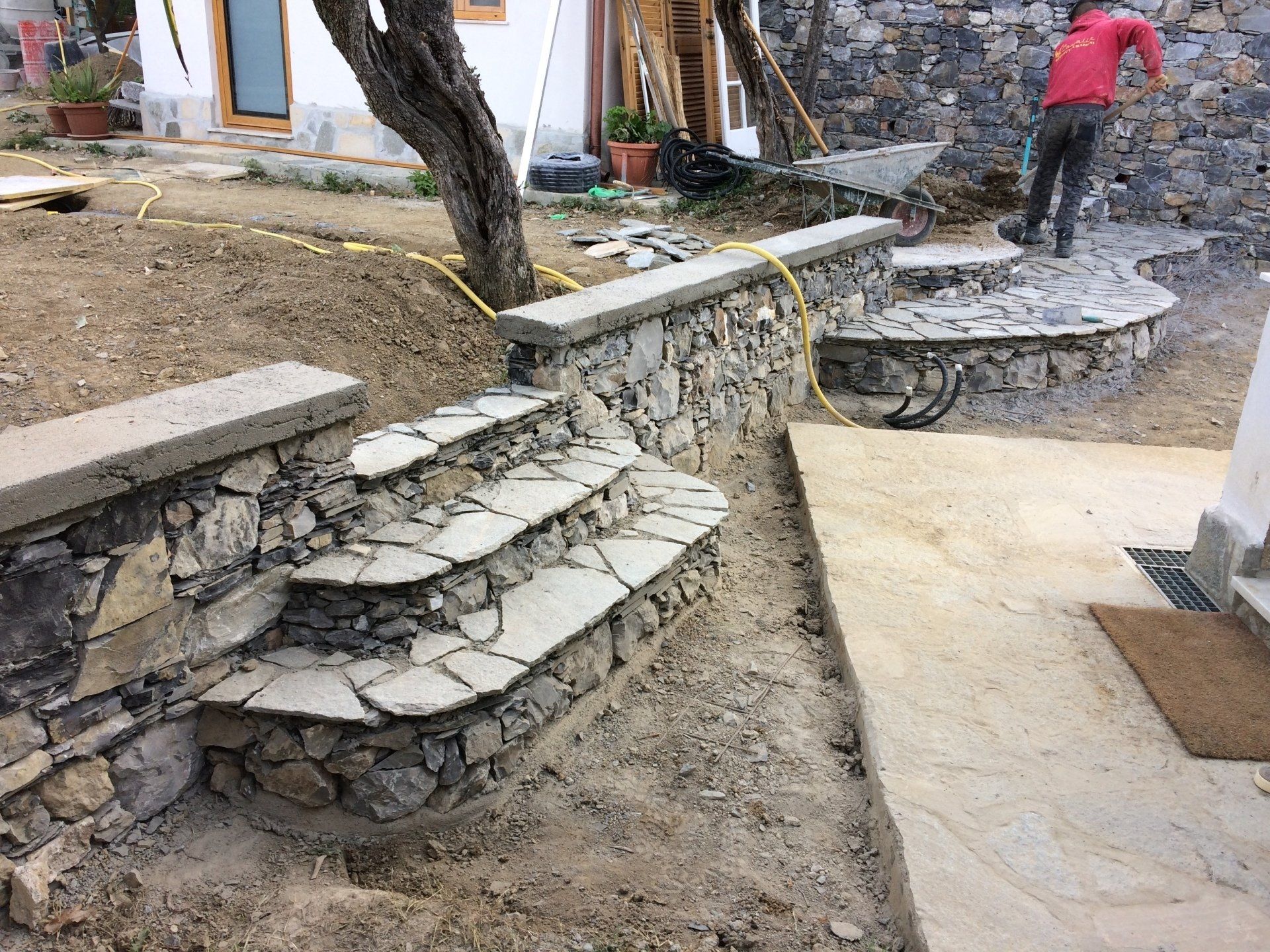 Gradini in pietra e muri di contenimento in costruzione in un cortile; un operaio usa una carriola.