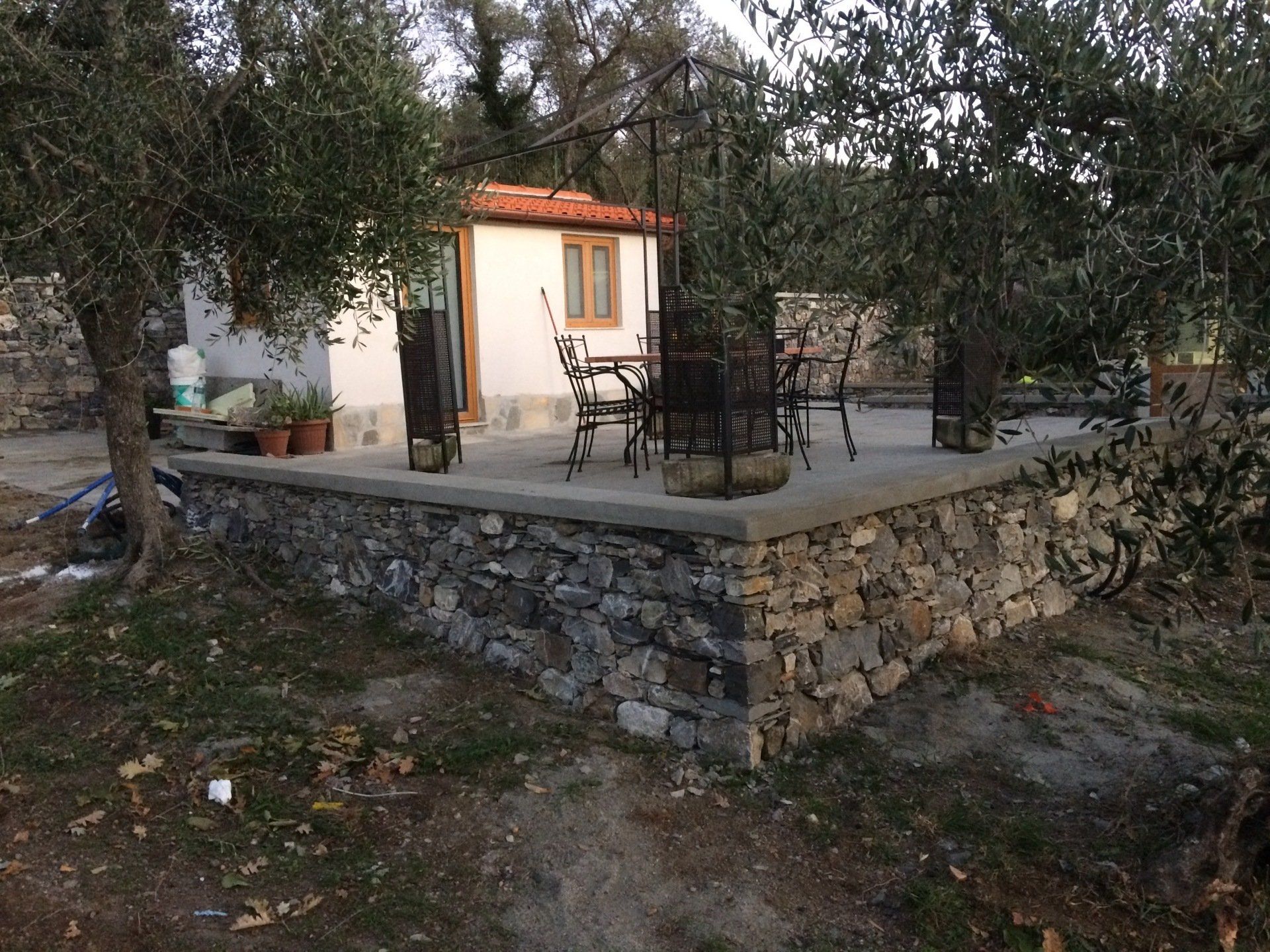 Patio in pietra con tavolo e sedie, adiacente a un piccolo edificio bianco con tetto marrone. Gli ulivi circondano il patio.
