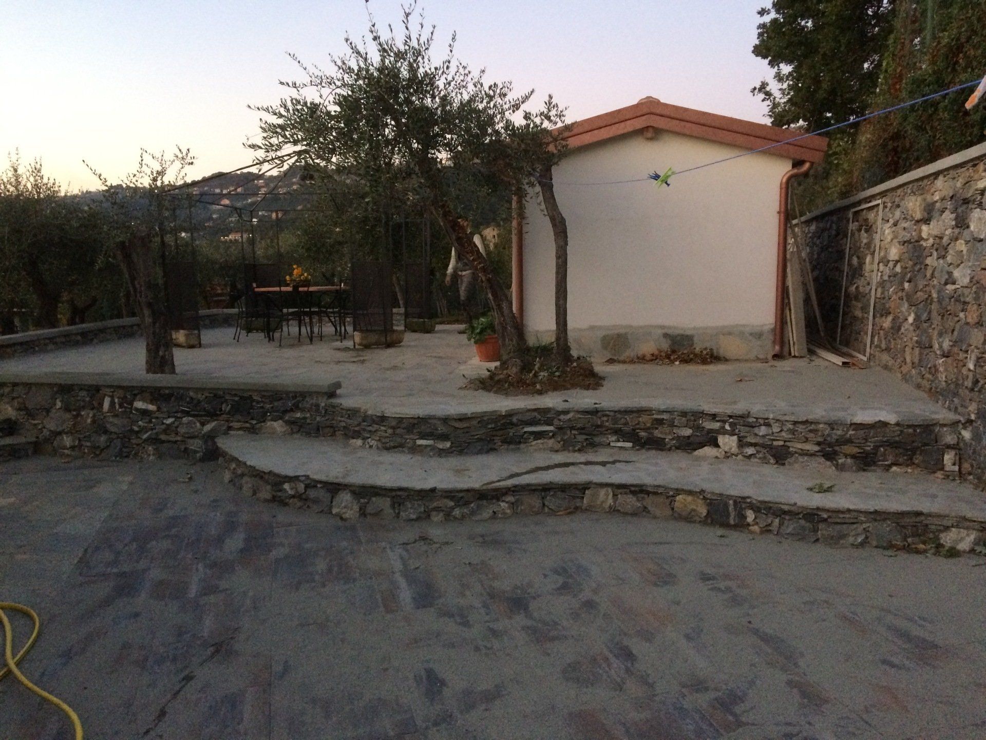 Patio in cemento con bordi in pietra, piccolo edificio e alberi. Luce serale.