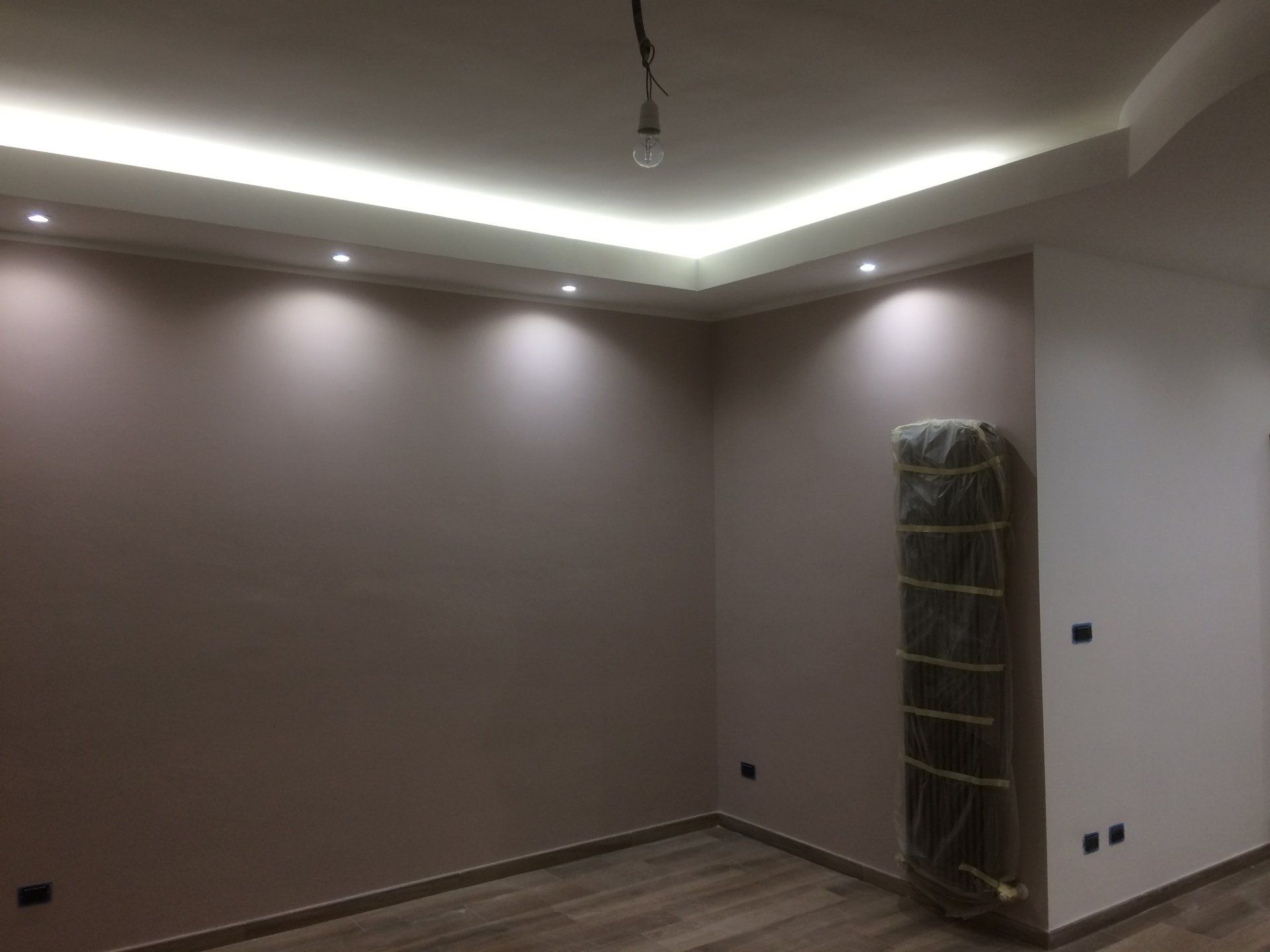 Stanza vuota con illuminazione a incasso e una striscia luminosa perimetrale sul soffitto. Pareti beige e pavimento in legno chiaro.