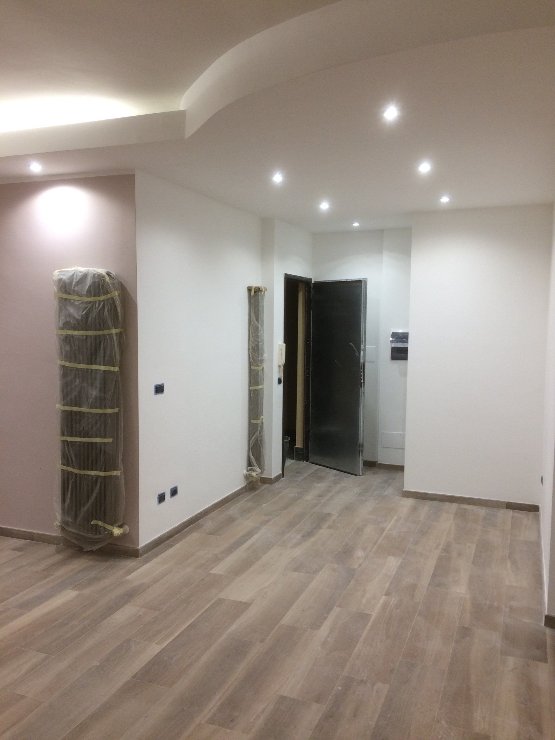 Stanza vuota con pavimenti in legno, pareti bianche, luci da incasso e una porta.