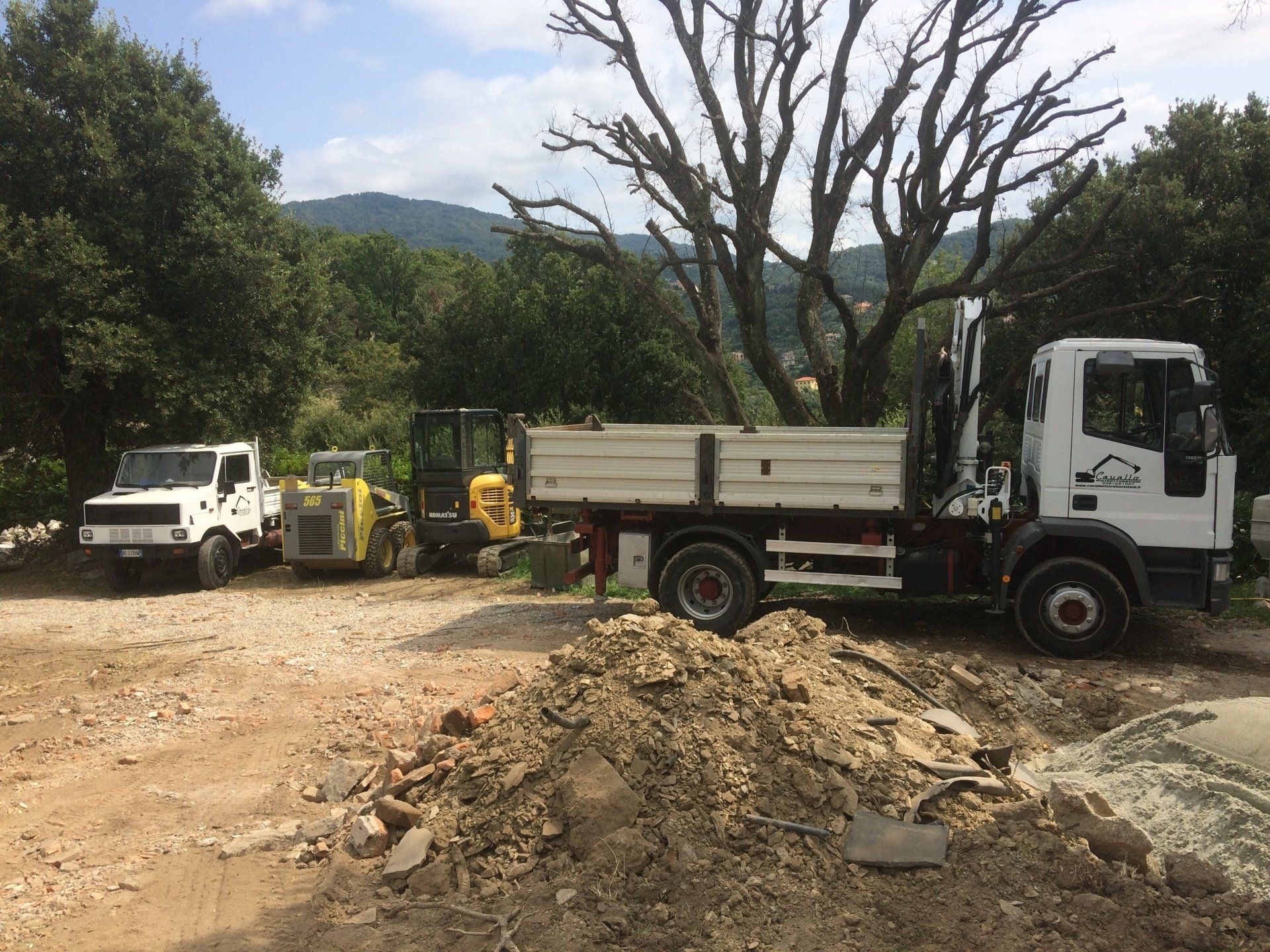 Un cantiere con un camion, un miniescavatore e un piccolo camion. Un cumulo di detriti in primo piano.