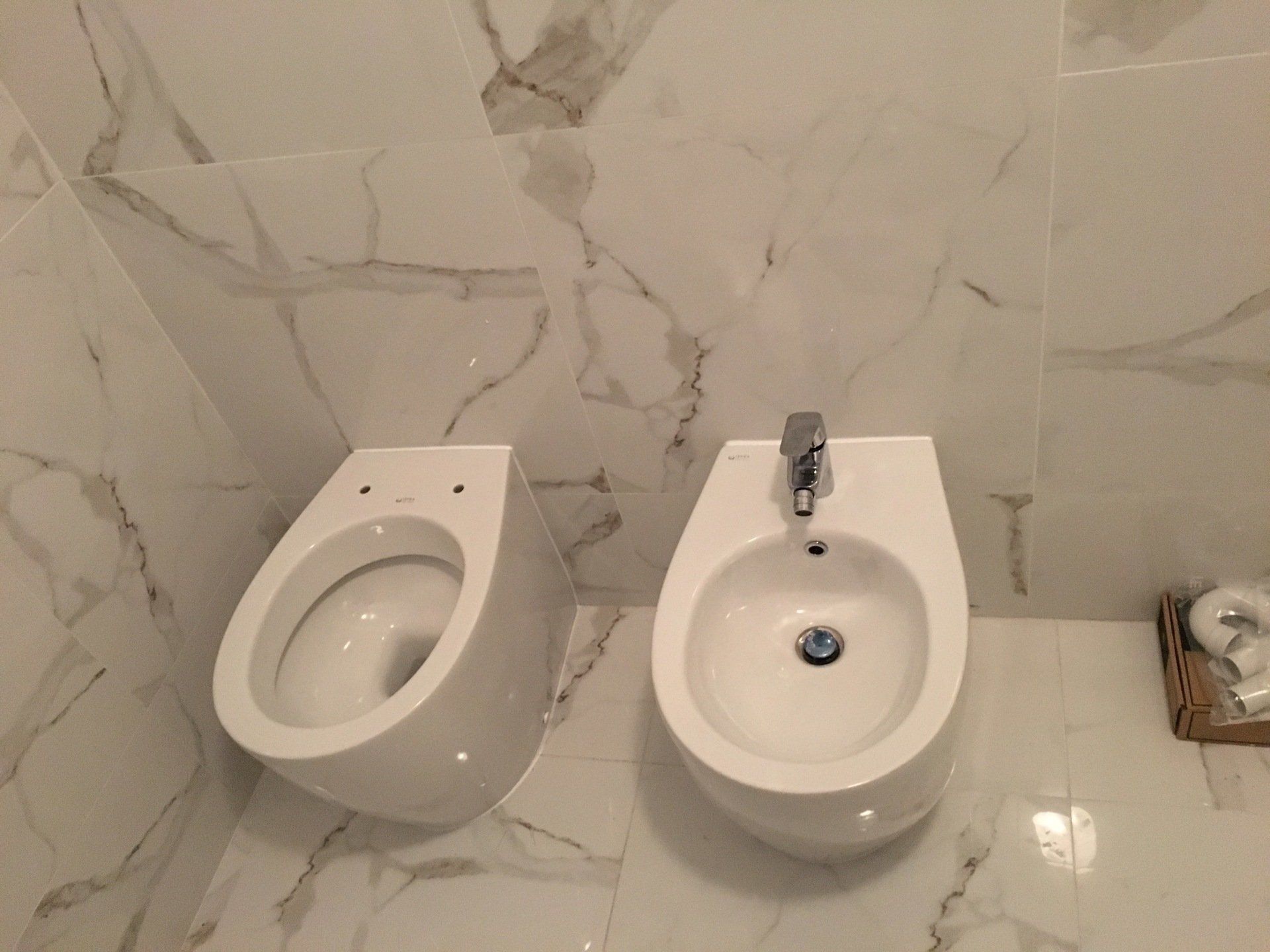 Sanitari per WC e bidet affiancati contro piastrelle a motivi marmorei.