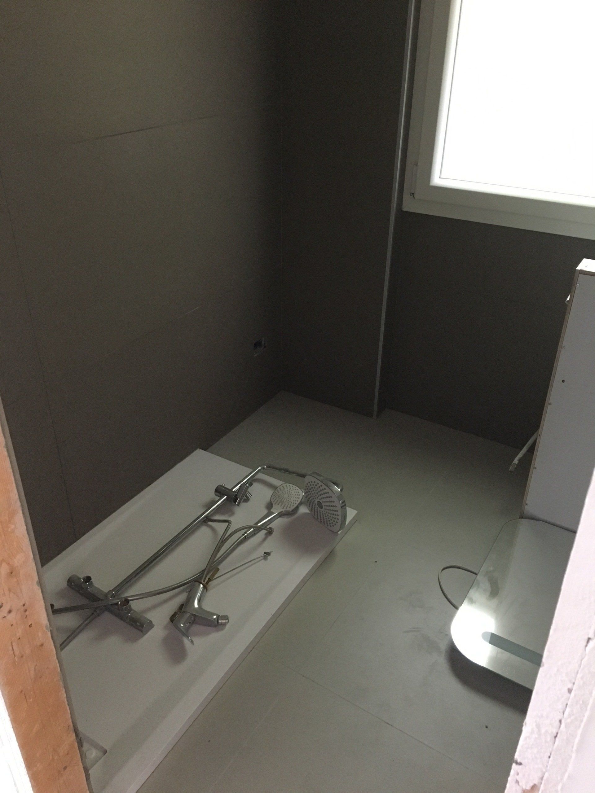Piccolo bagno con sanitari della doccia su una superficie bianca, pareti grigie e una finestra.