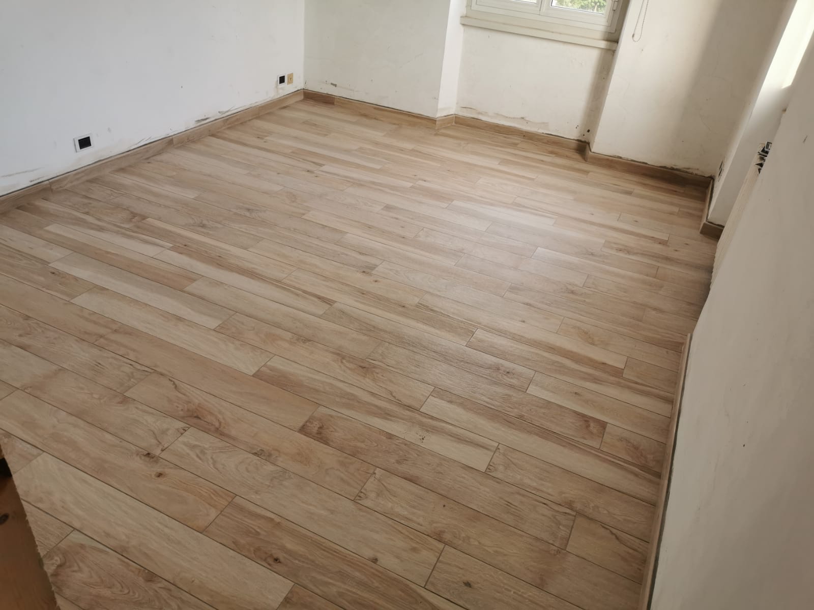 Stanza vuota con pavimento in legno chiaro. Sono visibili pareti bianche e una finestra.