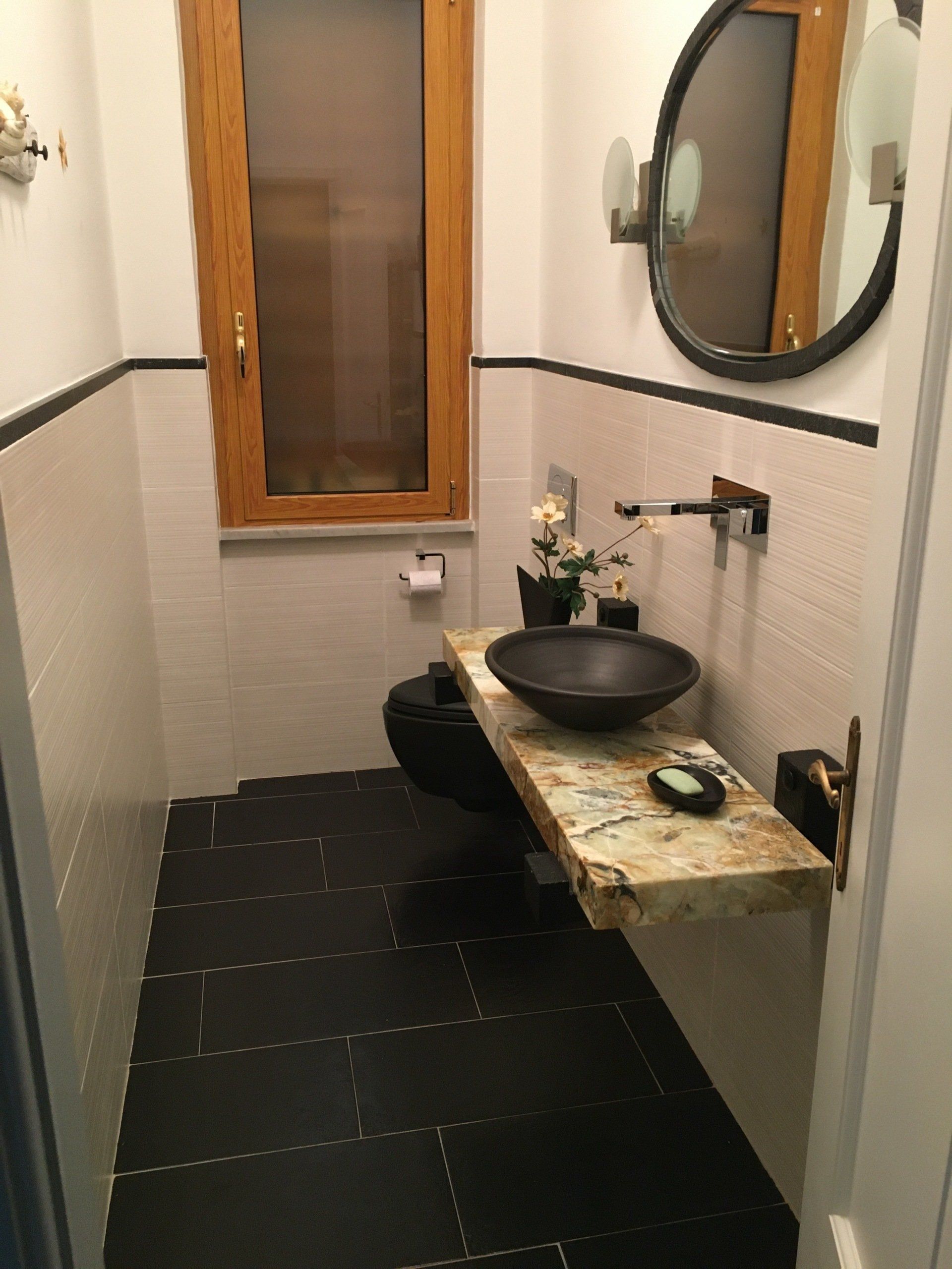 Piccolo bagno con water nero, lavandino su un piano in legno, finestra e pavimento in piastrelle nere.