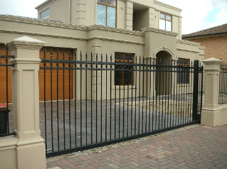 Black metal gate in front of a tan two-story house with a brick driveway.