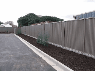 Long corrugated metal fence bordering asphalt, with mulch and small plants at its base.
