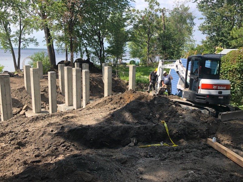 Chantier : Piliers en béton, mini-pelle, ouvriers, terre, arbres, eau en arrière-plan.