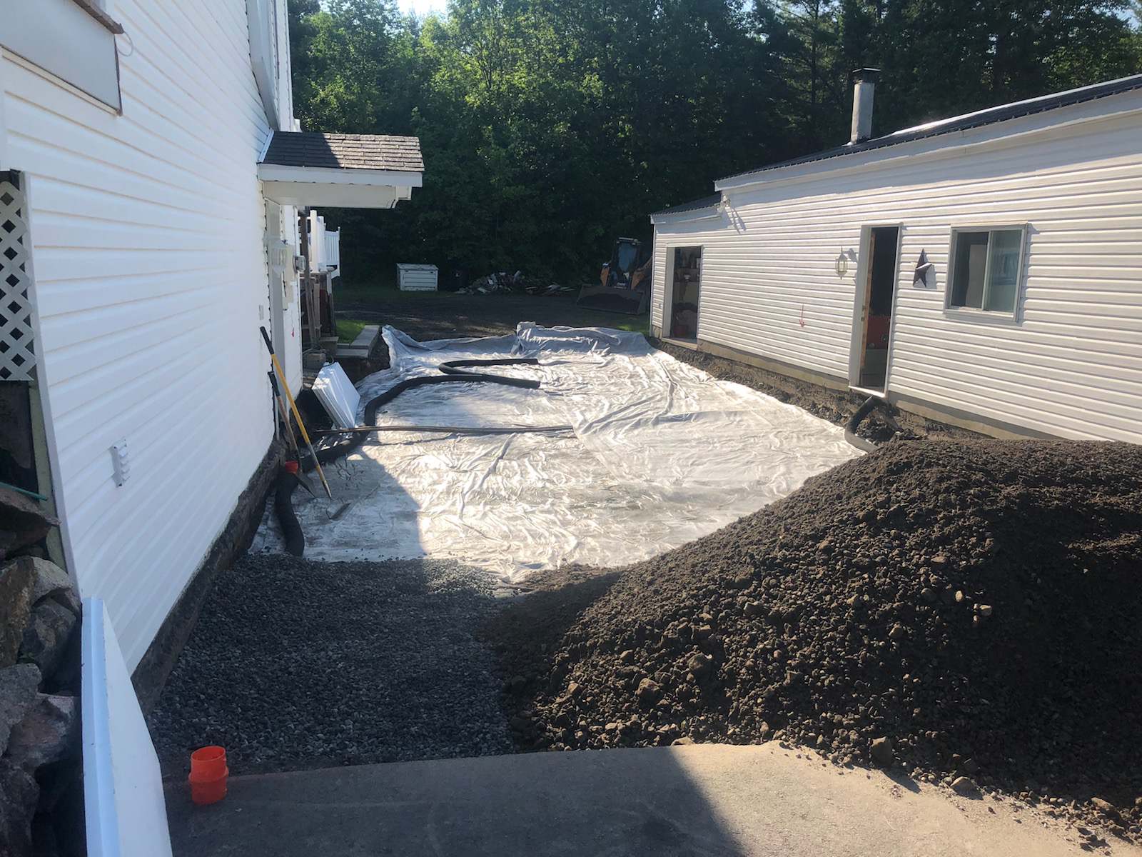 Zone recouverte de gravier entre une maison blanche et un garage. Présence de matériaux de construction et de débris.