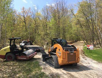 Chantier de construction avec engins lourds sur un chemin de terre : rouleau compresseur, chargeuse compacte, arbres en arrière-plan, journée ensoleillée.