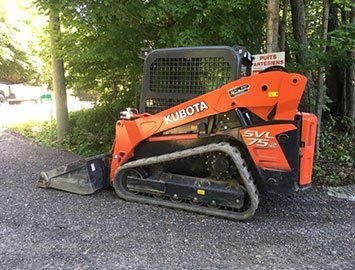 Chargeuse compacte à chenilles Kubota SVL75-2 orange sur du gravier, devant des arbres.