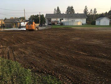 Un chantier de construction avec une chargeuse compacte orange sur un terrain en terre, un bâtiment en arrière-plan.