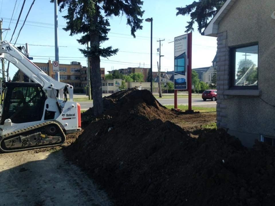 Une chargeuse compacte Bobcat à côté d'un tas de terre devant un bâtiment avec un panneau et une voiture rouge en arrière-plan.