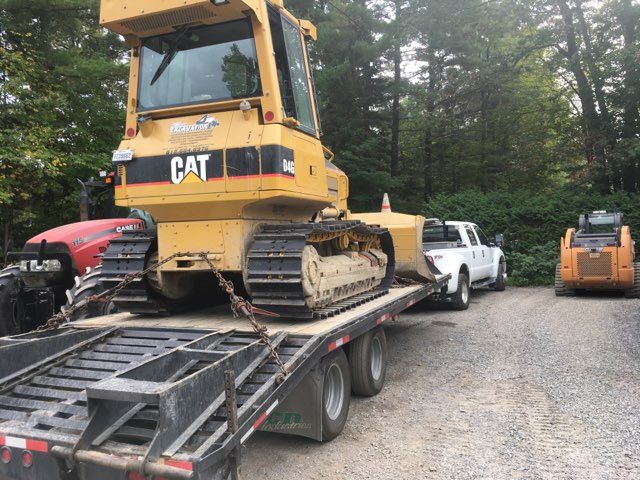 Un bulldozer Caterpillar jaune sur une remorque, chargé par un camion blanc, sur du gravier, avec une chargeuse compacte plus petite et un tracteur rouge en vue.