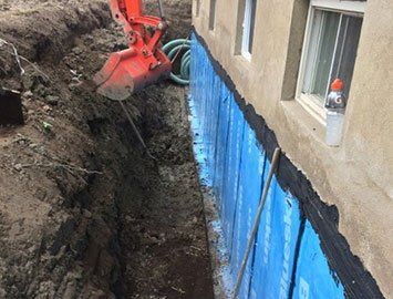 Une excavatrice creuse une tranchée à côté du bâtiment ; une membrane d'étanchéité bleue est installée.