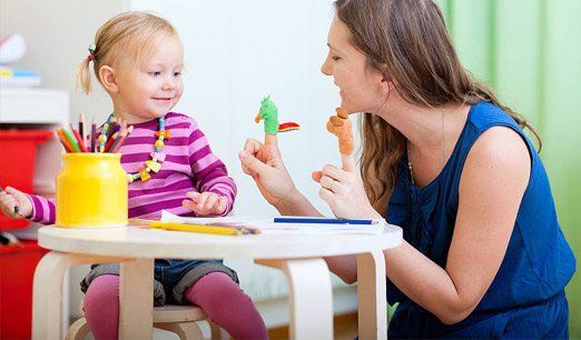 Woman and toddler at a white table, sharing toys and talking in a bright playroom.