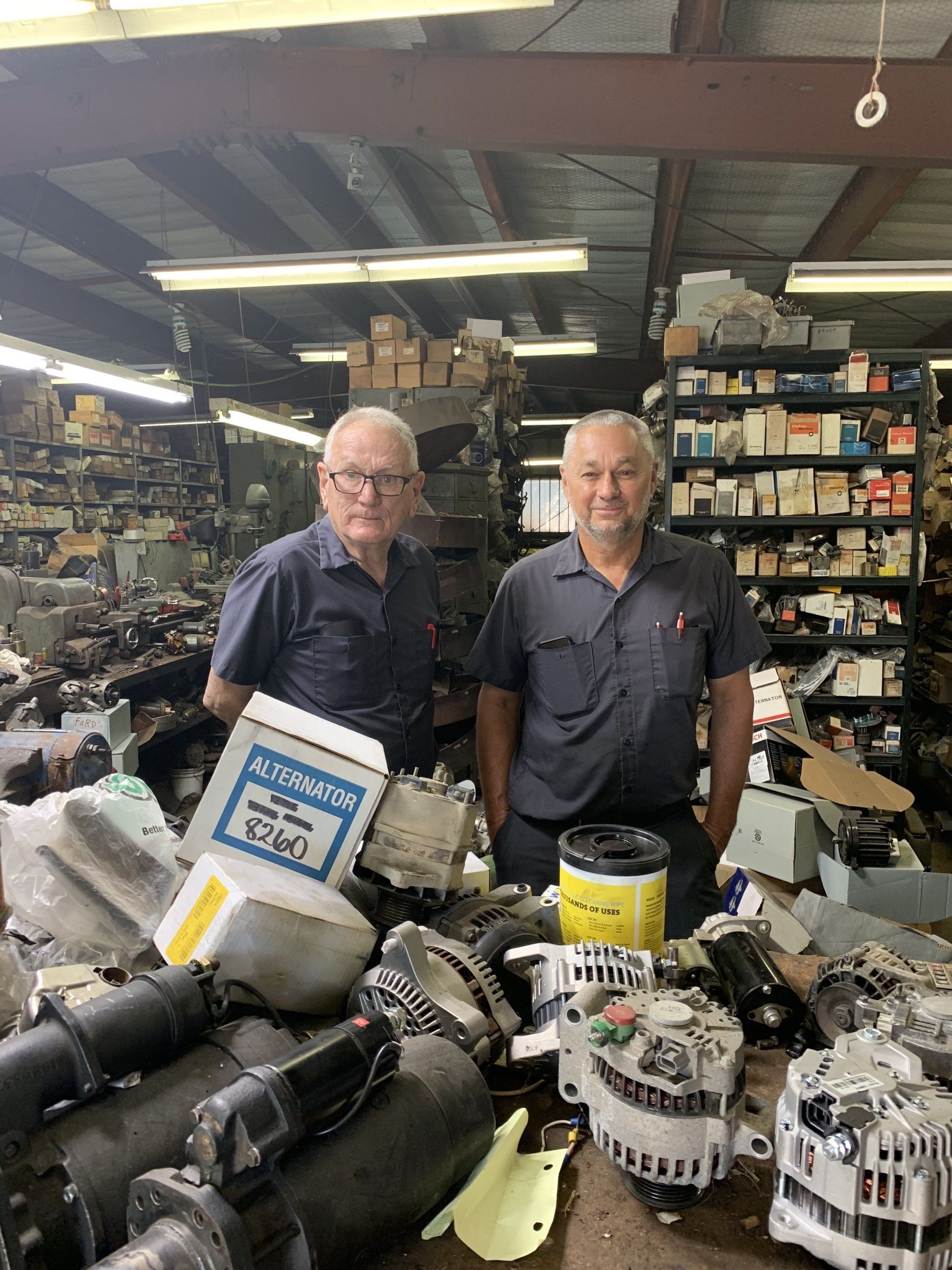 Two professionals stand in a cluttered auto parts warehouse surrounded by various mechanical components and shelves.