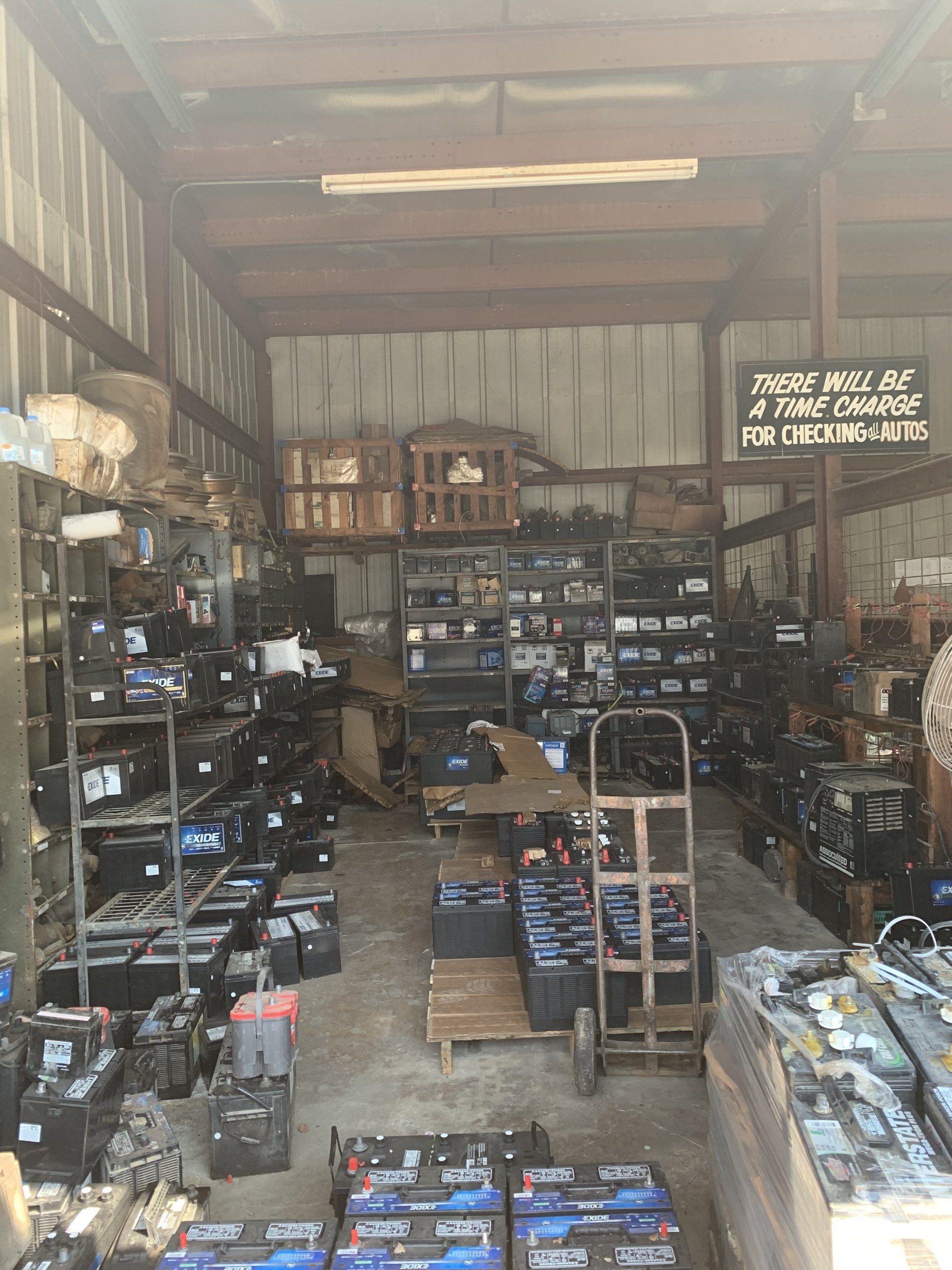 A cluttered industrial warehouse filled with shelves and pallets stacked with numerous automotive batteries.