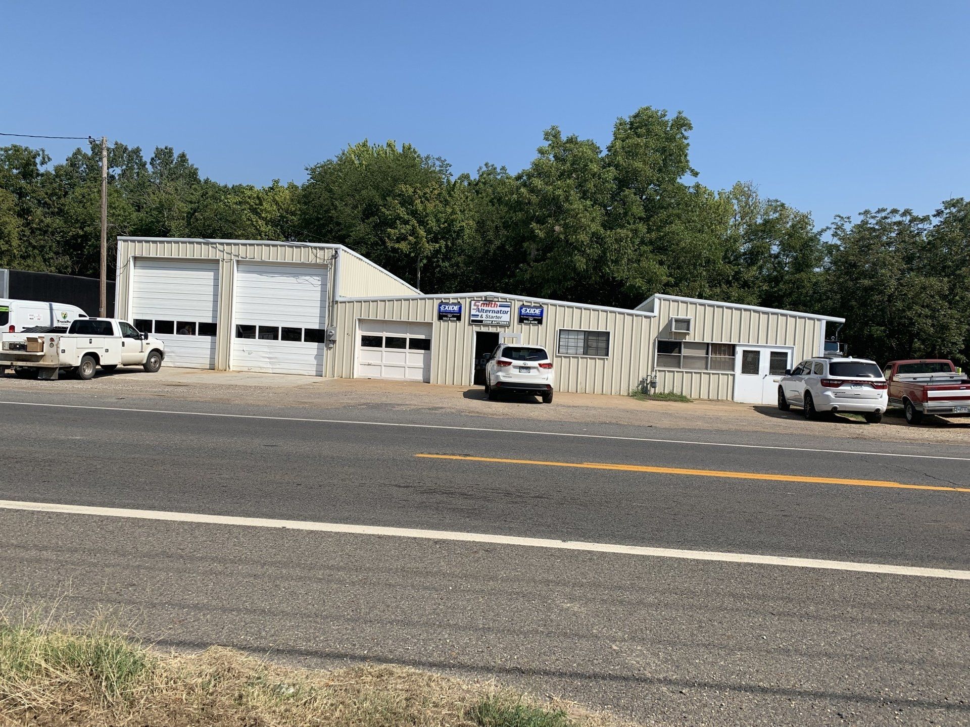 A one-story commercial auto repair shop with multiple garage bays, parked vehicles, and surrounding trees.