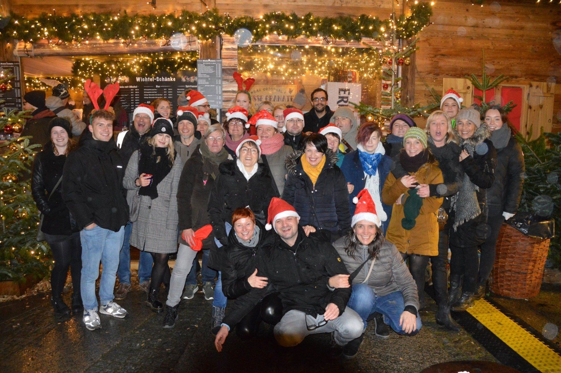 Eine Gruppe von Menschen mit Weihnachtsmannmützen posiert nachts vor einem warm beleuchteten Holzgebäude.
