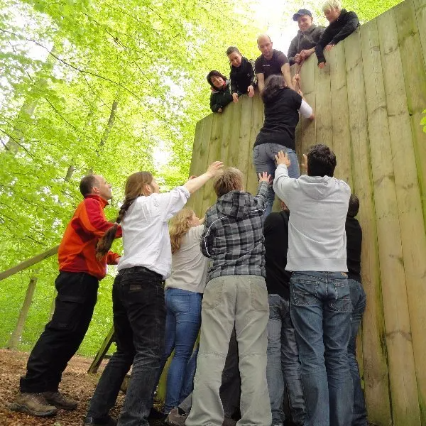 Eine Gruppe von Menschen hilft jemandem beim Erklimmen einer hohen Holzwand in einem grünen Waldgebiet.